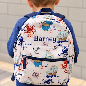 Peut inclure: Un sac à dos blanc avec un motif nautique composé de voiliers, de pieuvres rouges et bleues, d'ancres et de boussoles. Le prénom Barney est brodé en fil bleu foncé sur la poche avant, dotée d'une fermeture éclair et d'un liseré bleu foncé.