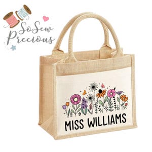 Può includere: Una borsa tote beige in iuta con un pannello frontale bianco con un colorato motivo floreale e il nome "MISS WILLIAMS" in nero. La borsa ha due manici beige e il testo "So Sew Precious" in alto a sinistra.