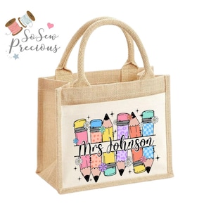 Può includere: Una borsa tote in iuta beige con una tasca frontale bianca con matite colorate e la scritta "Mrs Johnson". La borsa ha due manici beige ed è progettata per trasportare oggetti.