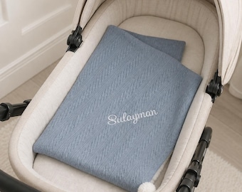 May include: A dusty blue knitted baby blanket with a cable knit texture and a small white pom-pom at the corner. The name Sulayman is embroidered in white cursive script in the center of the blanket, which is placed inside a beige baby carriage.