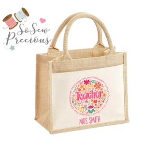 Può includere: Una borsa tote in iuta beige con un pannello frontale bianco con un design floreale e la parola "Teacher". Il design include parole come "Loving" e "Reliable". La borsa ha due manici e il testo "MRS SMITH".