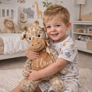 May include: A plush giraffe toy with a tan and brown spotted pattern, featuring a light brown belly patch embroidered with the name Uswa in white script. The stuffed animal has a soft texture, a small tan ribbon bow at the neck, and dark eyes.
