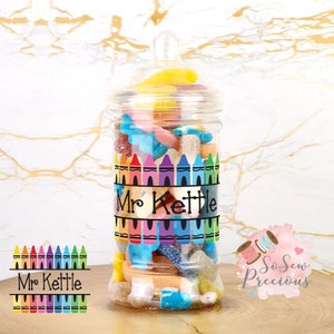 Peut inclure: Un bocal en verre transparent rempli de bonbons colorés. Le bocal porte une étiquette avec un arc-en-ciel de crayons et le texte "Mr Kettle".