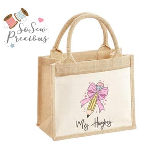 Può includere: Una piccola borsa tote in iuta beige con un pannello frontale bianco. La parte anteriore presenta un disegno di matita gialla con un fiocco rosa e il nome "Mrs. Hughes" in corsivo nero. La borsa ha due manici beige e il testo "So Sew Precious".