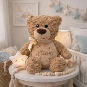 Peut inclure: Un ours en peluche marron avec un ruban crème et le nom "Liam" brodé sur la poitrine. L'ours est entouré d'une veilleuse en forme d'étoile et de perles en bois. Ambiance de chambre d'enfant.