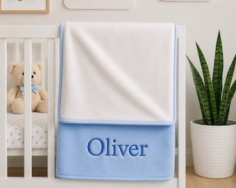 May include: A soft, rectangular baby blanket featuring a white top section and a solid light blue bottom section. The name Oliver is embroidered in dark blue thread across the light blue portion of the blanket, which is draped over a white crib rail.