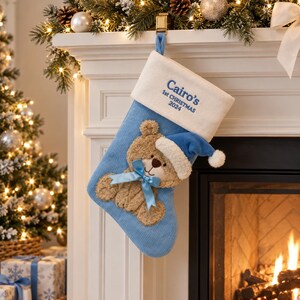May include: A light blue corduroy Christmas stocking with a white cuff featuring the embroidered text "Cairo's 1st CHRISTMAS 2024". The stocking displays a plush tan teddy bear wearing a blue hat and a light blue satin ribbon bow at its neck.