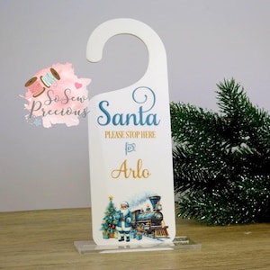 Può includere: Appendiabiti bianco con parte superiore curva, con la scritta "Santa PLEASE STOP HERE for Arlo" in blu e oro. Un'illustrazione festosa di Babbo Natale, un albero di Natale e un treno si trova in basso. L'appendiabiti è su un supporto trasparente.