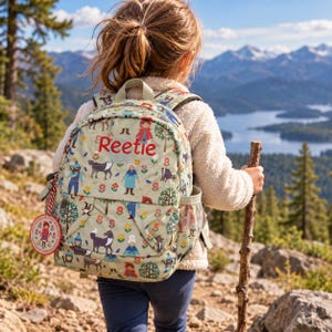 May include: A light green backpack featuring a repeating pattern of characters, trees, and flowers, with the name Reetie embroidered in red thread on the front pocket. A round tag with a character illustration hangs from the side zipper.