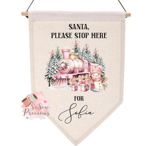 Può includere: Striscione natalizio beige con un treno rosa, un pupazzo di neve e dei regali. Il testo recita "SANTA, PLEASE STOP HERE" e "FOR Sofia". Lo striscione è sospeso da un tassello di legno.