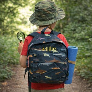 May include: A navy blue backpack with a shark pattern and the name "Millie" in yellow. The backpack is worn by a person wearing a camouflage hat and red shirt, carrying a magnifying glass and a blue yoga mat.