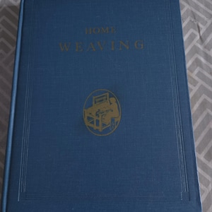 Peut inclure: Un livre bleu à couverture rigide intitulé "HOME WEAVING" en lettres dorées. La couverture présente une illustration dorée d'un métier à tisser. Le livre a un aspect texturé et est probablement un guide du tissage à domicile.