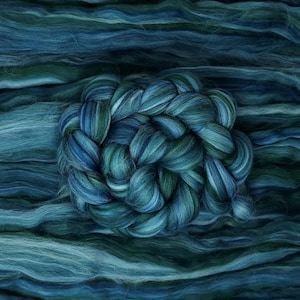 May include: A close-up of a skein of hand-dyed fiber in shades of teal, blue, and green. The fiber is arranged in a circular braid, surrounded by loose, wavy strands of the same colors. The texture appears soft and fluffy, ideal for crafting.