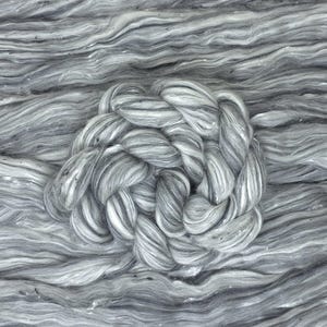Puede incluir: Primer plano de hilo gris y blanco. El hilo está dispuesto en forma de nudo trenzado, rodeado de hebras sueltas. La paleta de colores es principalmente gris y blanco, con sutiles variaciones de tono y textura. El hilo parece suave y esponjoso, adecuado para tejer u otras manualidades.