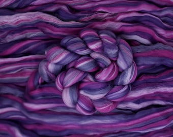 Puede incluir: Primer plano de un paquete de vellón de lana púrpura, rosa y blanca. El vellón está dispuesto en un patrón de remolino, creando una textura visualmente atractiva.