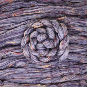 Puede incluir: Primer plano de un ovillo de lana teñida a mano en tonos morados, con motas rojas, naranjas y amarillas. La lana está dispuesta en espiral, con hebras sueltas alrededor. Ideal para proyectos de punto y ganchillo.