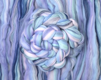 May include: Close-up of a bundle of blue, white, and lavender wool roving. A braided section of the roving is in the center, surrounded by loose strands. The colors blend together, creating a soft, textured appearance, ideal for fiber art projects.