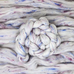 Puede incluir: Primer plano de lana de mecha blanca con motas azules y moradas. Una sección de la lana está trenzada en forma circular. La lana tiene un aspecto suave y texturizado, ideal para proyectos de manualidades.