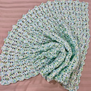 Peut inclure: Une couverture au crochet faite à la main, dans des tons de blanc, bleu clair et vert. La couverture a un bord festonné et un motif ajouré texturé. La couverture est pliée, révélant le travail de couture complexe.