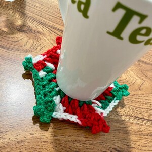 May include: A white ceramic mug rests on a handmade crochet coaster. The coaster features a festive color scheme of red, green, and white. The mug has green text on it. The coaster is square-shaped and appears to be made of yarn.