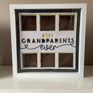 Puede incluir: Marco de fotos blanco con seis aberturas. El texto "BEST GRANDPARENTS ever" está en oro y azul marino. La pantalla está sobre una superficie blanca.