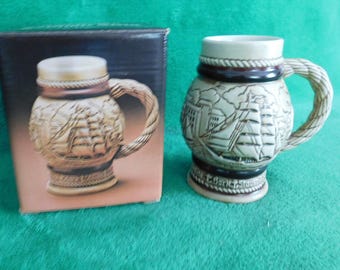 May include: A beige ceramic beer stein with a nautical design featuring a ship and buildings. The handle is rope-like, and the stein is next to its box. The background is green.