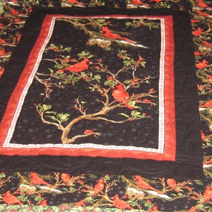 May include: A black and red quilt with a central panel featuring a cardinal bird perched on a branch with red berries. The quilt is surrounded by a black border and a red and white border.