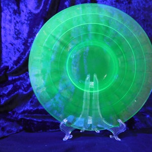 May include: A bright green glass plate with a concentric circle design, displayed on a clear plastic stand. The plate's colour is a glowing green. The plate is likely vintage or antique.