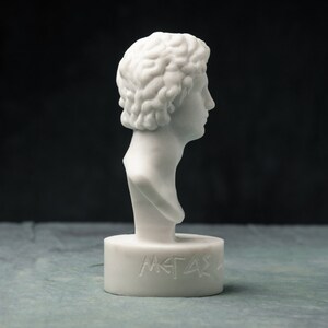May include: A white marble bust of a man with curly hair, seen in profile. The bust is mounted on a cylindrical base with engraved text. The sculpture has a classical aesthetic and is set against a dark background.