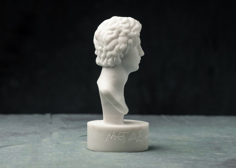 May include: A white marble bust of a man with curly hair, seen in profile. The bust is mounted on a cylindrical base with engraved text. The sculpture has a classical aesthetic and is set against a dark background.