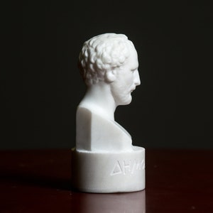 Puede incluir: Busto de m&aacute;rmol blanco que representa la cabeza y los hombros de un hombre. El busto est&aacute; sobre un pedestal con la palabra "&Delta;&Eta;&Mu;&Omicron;" inscrita en la parte delantera.