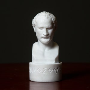 Puede incluir: Busto de m&aacute;rmol blanco de un hombre con el pelo corto y barba. El busto est&aacute; sobre una base blanca con letras griegas.