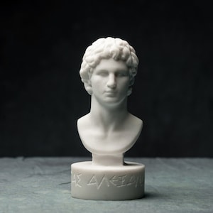 May include: A white marble bust of a man with curly hair and a serene expression. The bust is mounted on a cylindrical base with engraved text. The sculpture is set against a dark background.