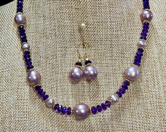 May include: A necklace and earring set featuring large, light purple pearls and smaller, faceted purple beads. The necklace has gold-colored accents. The earrings have matching purple pearl drops. The jewelry is displayed on a tan mannequin.