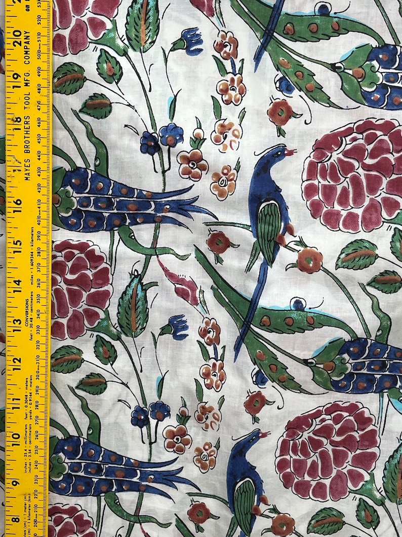 May include: White fabric with a repeating pattern of stylized peacocks, flowers, and leaves. The peacocks are blue, green, and gold. The flowers are red, orange, and yellow. The leaves are green.