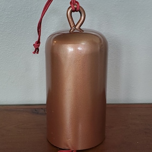 Może przedstawiać: Dzwonek w kolorze miedzi z czerwonym sznurkowym uchwytem. Dzwonek jest gładki i błyszczący.