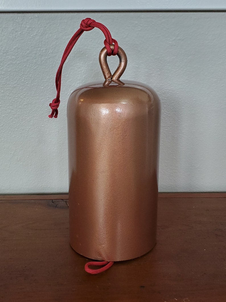 Może przedstawiać: Dzwonek w kolorze miedzi z czerwonym sznurkowym uchwytem. Dzwonek jest gładki i błyszczący.