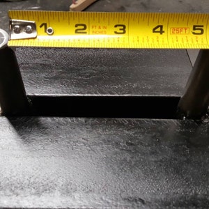 May include: Close-up of a black metal weightlifting stand with two metal posts. A yellow tape measure is placed across the top of the stand, showing measurements in inches and feet.