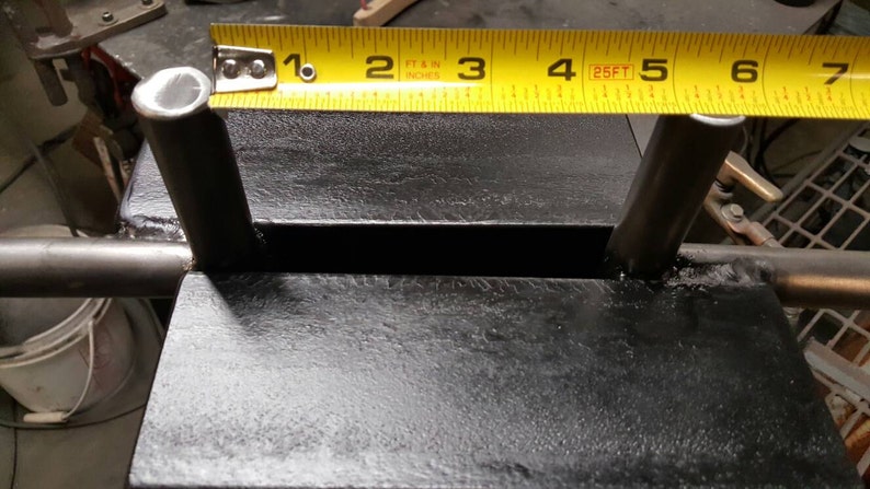 May include: Close-up of a black metal weightlifting stand with two metal posts. A yellow tape measure is placed across the top of the stand, showing measurements in inches and feet.