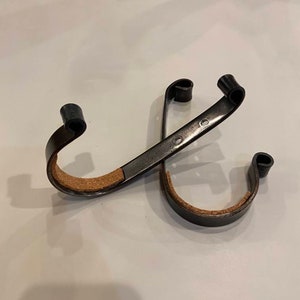 May include: Two black metal hooks with brown leather straps. The hooks are designed to hang items on a wall or door.