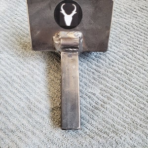 May include: A metal T-shaped tool with a flat top and a sticker of a white deer head on a black circle. The tool is made of metal and has a rough, unfinished look.