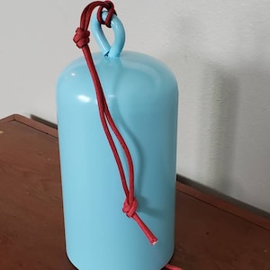 Może przedstawiać: Dzwonek metalowy w jasnoniebieskim kolorze z czerwonym sznurkiem jako uchwytem. Dzwonek ma kształt cylindryczny i gładką powierzchnię.