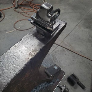 May include: A metal anvil with a metal object on top. The object is a square shape with a loop on the side. The anvil is sitting on a metal table.