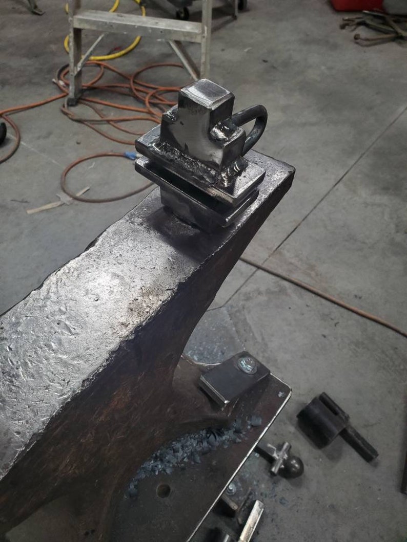 May include: A metal anvil with a metal object on top. The object is a square shape with a loop on the side. The anvil is sitting on a metal table.