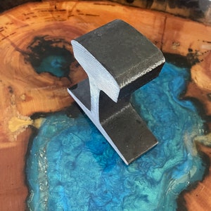 May include: A black metal railroad track section, shaped like an 'I', is displayed on a wooden surface with a blue resin inlay.