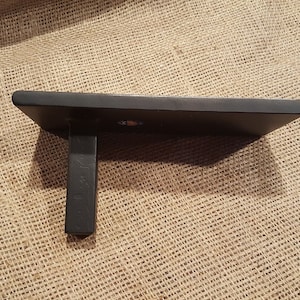 May include: Black wooden shelf with a single bracket. The shelf is rectangular and has a smooth surface. The bracket is attached to the underside of the shelf and is also made of wood.