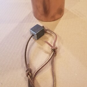 Może przedstawiać: Brązowy sznurek sk&oacute;rzany z kwadratowym ciężarkiem metalowym przymocowanym do jednego końca. Sznurek jest zawiązany w supeł.