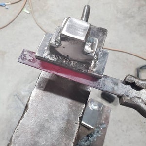 May include: A metal anvil with a square metal block attached to it. A red-hot metal rod is being hammered on the anvil.