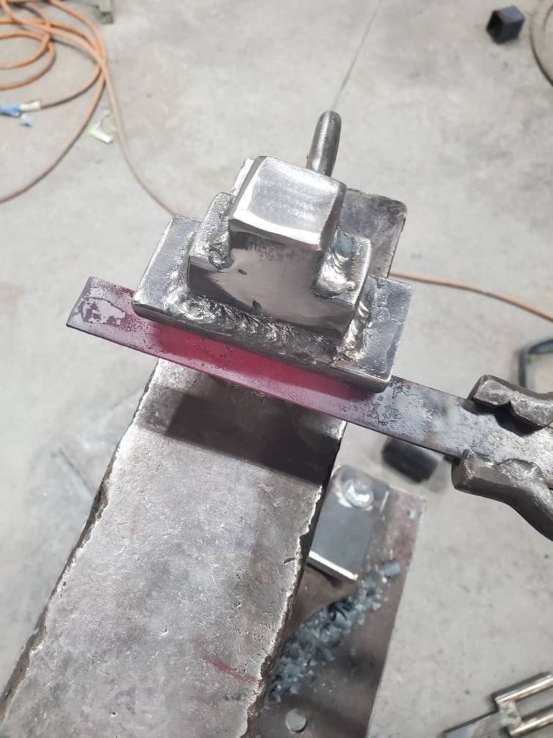 May include: A metal anvil with a square metal block attached to it. A red-hot metal rod is being hammered on the anvil.