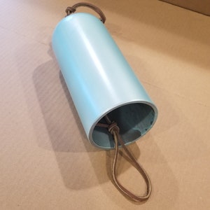 Może przedstawiać: Dzwonek wietrzny w kształcie cylindra w jasnoniebieskim kolorze z brązową sk&oacute;rzaną pętlą. Dzwonek jest zawieszony na pętli.